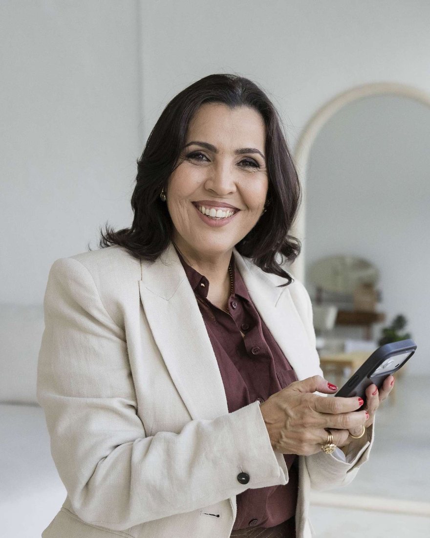 Tatiana Teixeira sorrindo, de blazer claro, com o celular na mão